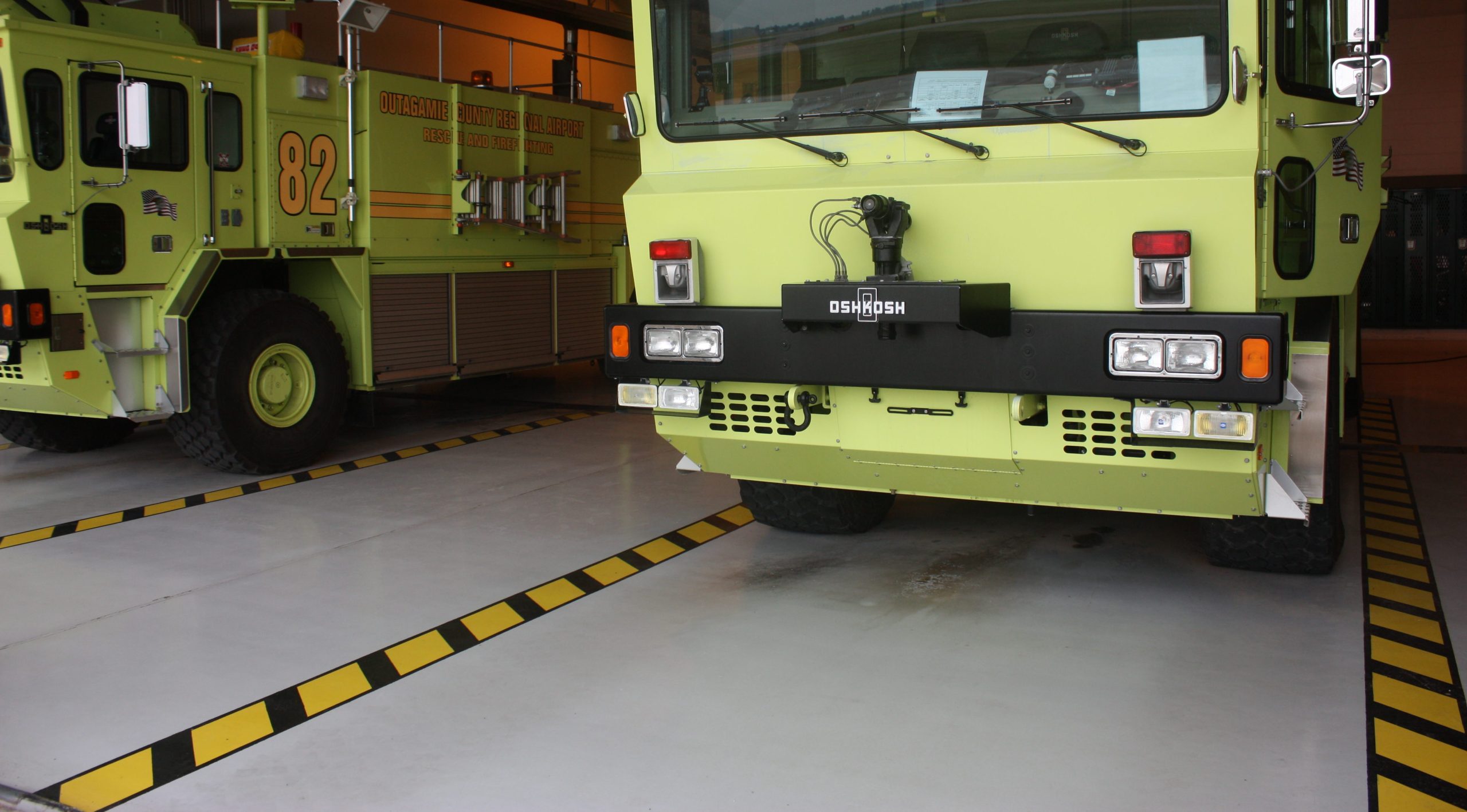 Fire truck parked inside a fire station, featuring a bright green design, equipped with emergency lights and firefighting tools. The vehicle is positioned on a marked floor with black and yellow safety stripes, highlighting its readiness for emergency response.