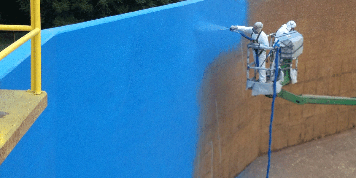A painter in a white suit uses a spray gun to apply a vibrant blue coat of paint to a large wall, showcasing professional painting techniques and equipment.