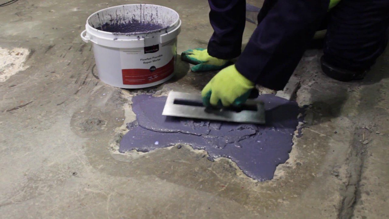 Worker applying purple flowable repair mortar to a concrete floor using a trowel, with a bucket of the product nearby.