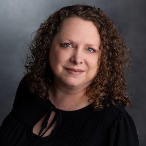 Professional headshot of a woman with curly hair, wearing a black blouse, smiling confidently against a gray background.