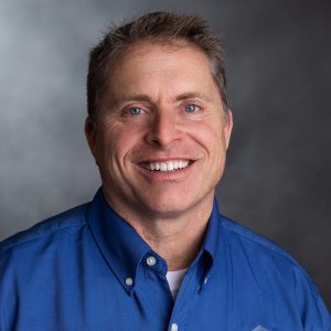 Smiling man in a blue shirt with short hair, posing against a gray background, representing a professional headshot for business or personal branding.
