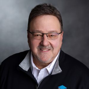 Professional headshot of a middle-aged man with glasses, smiling, wearing a black jacket with a logo, against a neutral gray background.
