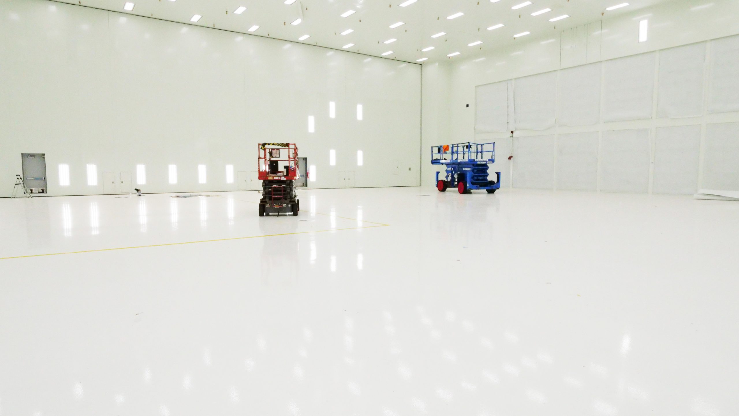 Spacious industrial facility interior featuring a glossy white floor and two elevated work platforms, one red and one blue, against a backdrop of bright white walls and ample natural light.
