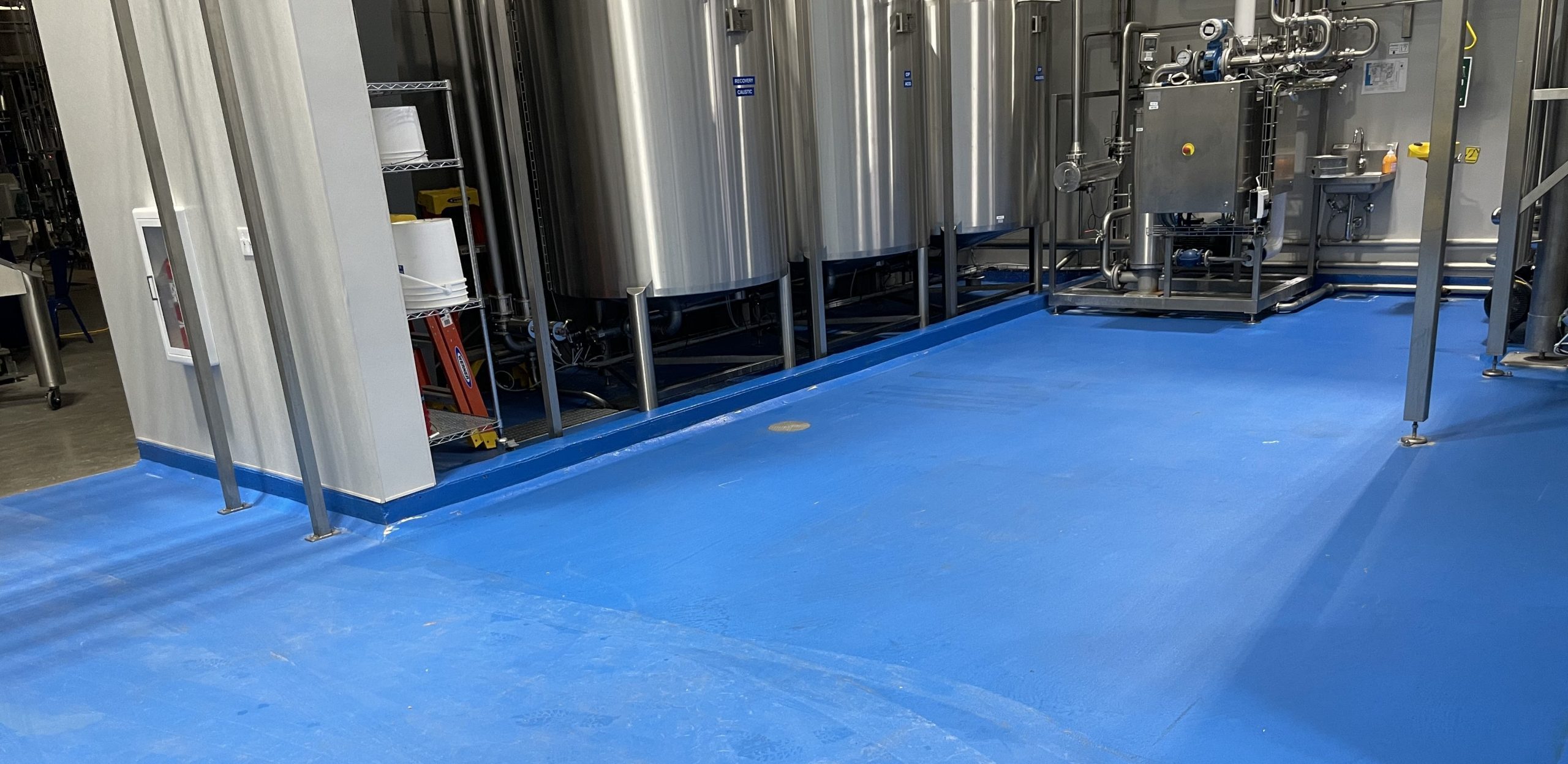 Industrial brewing facility with stainless steel fermentation tanks and a blue epoxy floor, showcasing a clean and organized workspace.