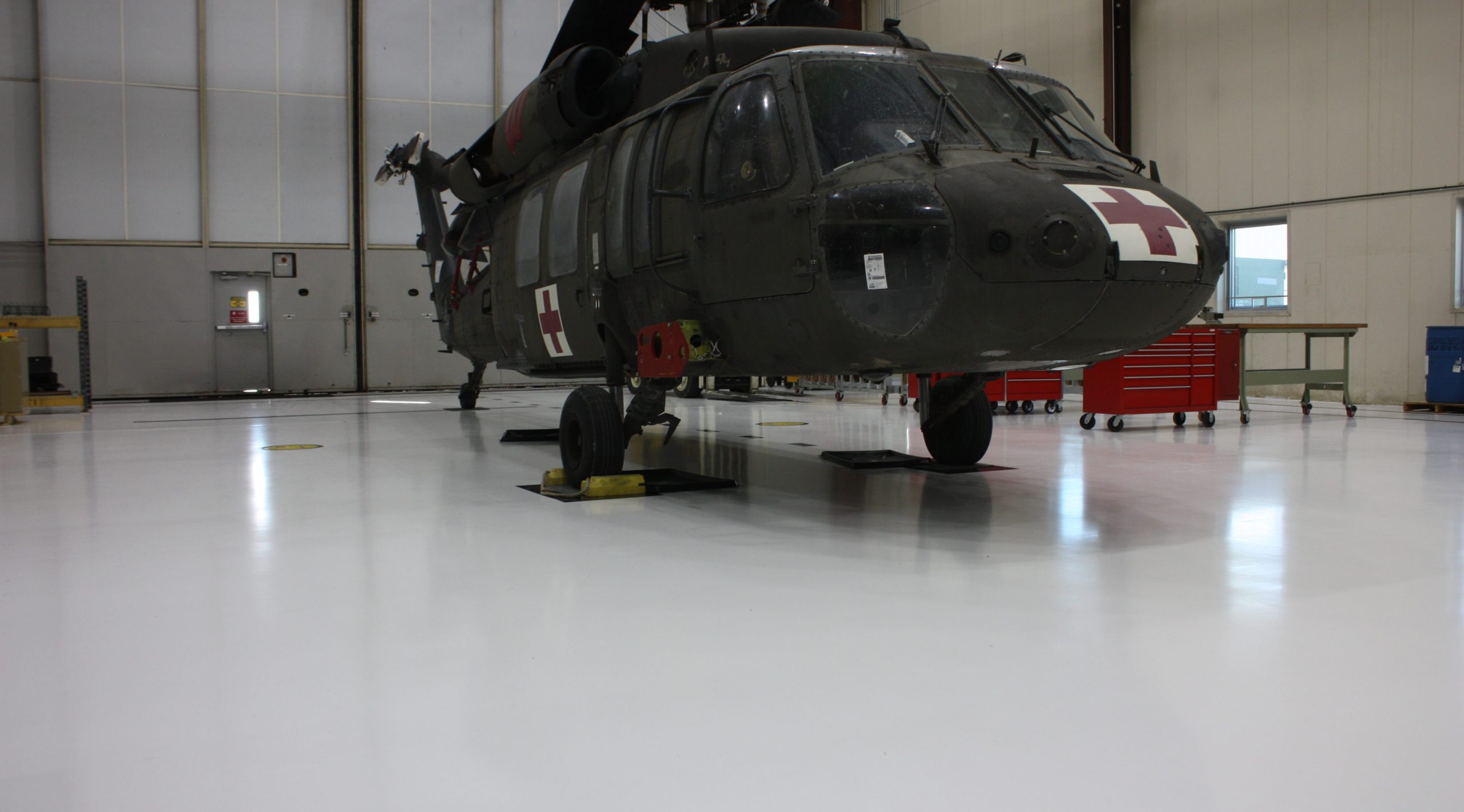 Black Hawk helicopter in a maintenance hangar, featuring a red cross emblem, positioned on support stands with a clean, white floor and various tools and equipment visible in the background.