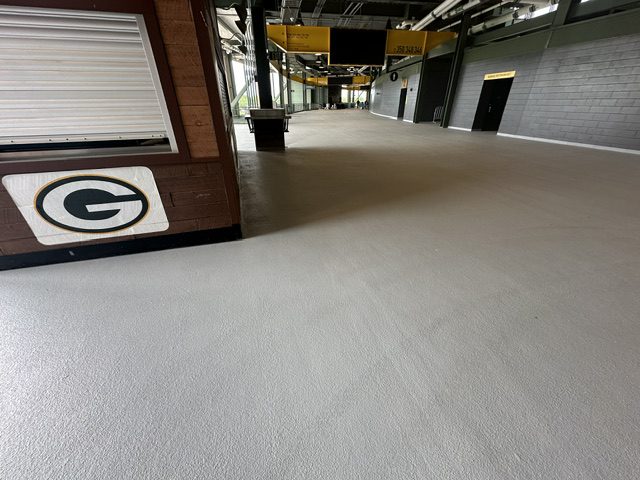 Interior view of a sports venue featuring a smooth, gray floor, with a concession stand displaying the Green Bay Packers logo on the left. The space is well-lit and includes various access points along the walls.