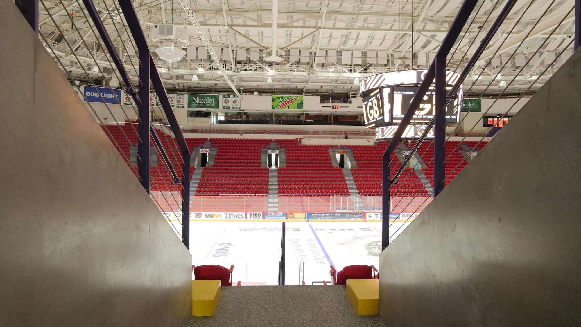 View from the entrance of a hockey arena, showcasing the empty ice rink and red seating. The overhead scoreboard displays the game time, while advertisements are visible on the walls.