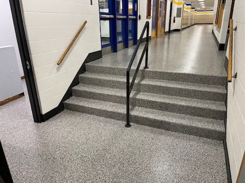 Interior view of a school hallway featuring a set of gray stairs with a black handrail, leading to an upper level. The floor has a speckled finish, and double doors are visible in the background.