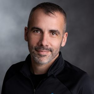 Professional headshot of a man with short gray hair and a beard, wearing a black jacket, set against a gray background.