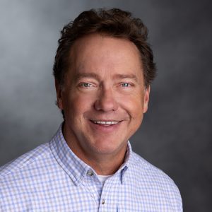 Smiling middle-aged man with short brown hair wearing a checkered shirt, posing against a neutral gray background.