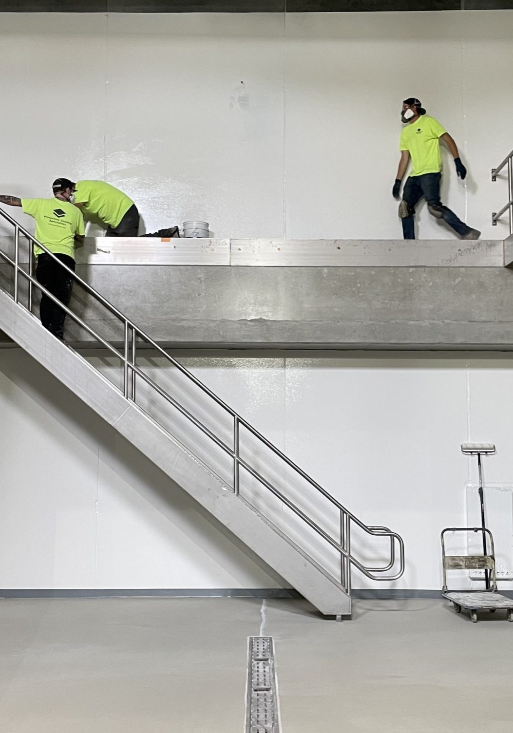 Workers in bright green safety shirts are performing tasks on a multi-level platform with a metal staircase in an industrial setting.