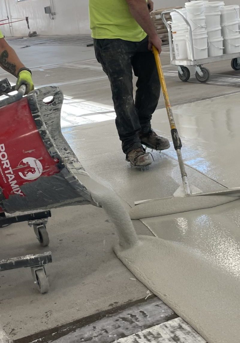 Two workers in yellow shirts are using a concrete mixer and a trowel to pour and smooth a fresh layer of concrete on a floor in a large industrial space. Buckets of paint or materials are visible in the background, indicating ongoing construction or renovation work.