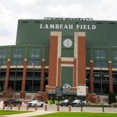 Exterior view of Lambeau Field, home of the Green Bay Packers, showcasing the iconic green and brick structure, American flag, and surrounding parking area.